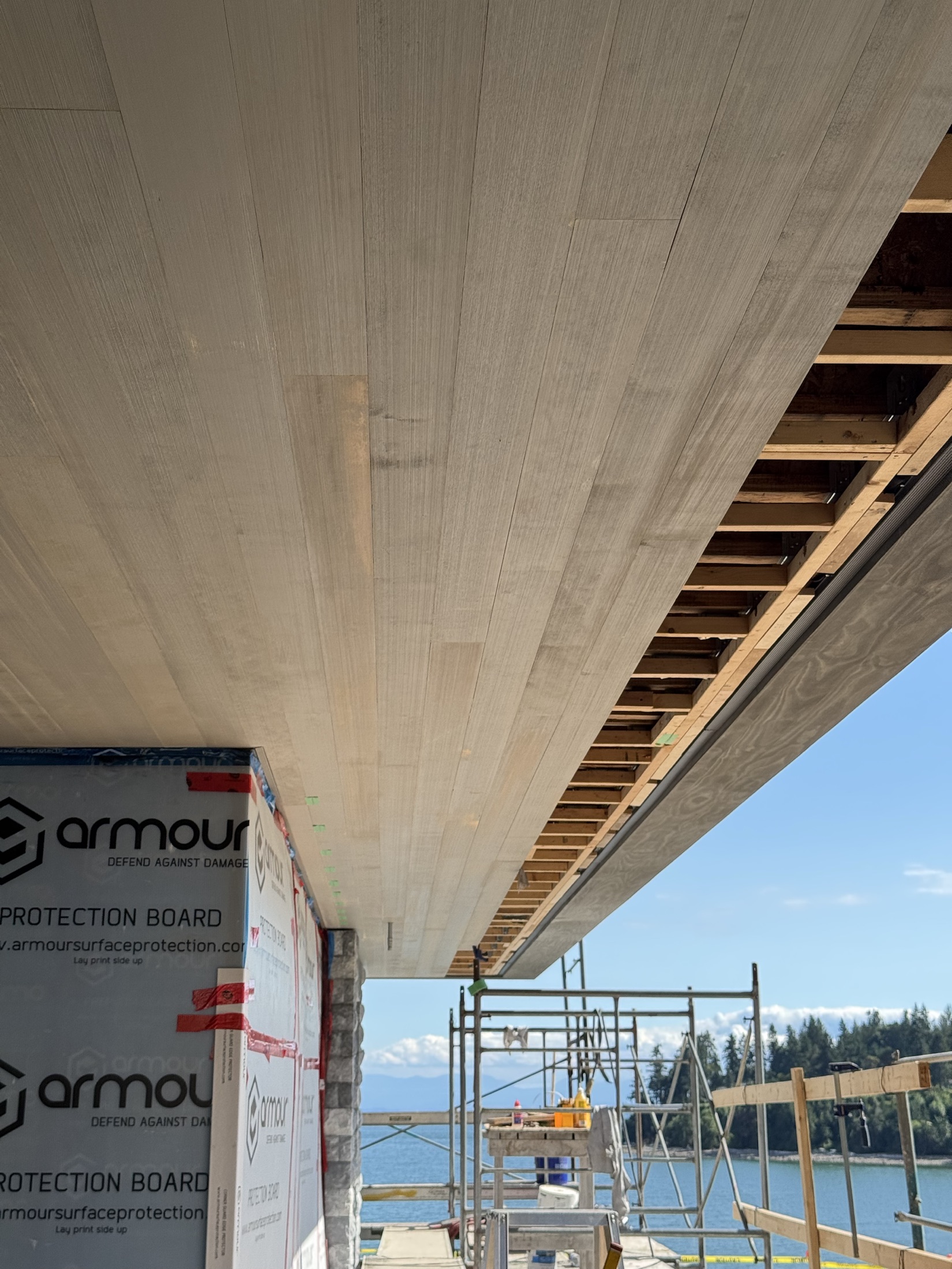 Cove Beach exterior hemlock soffit mid-install — finished hemlock on one side, exposed framing on the other, ocean beyond, Sechelt